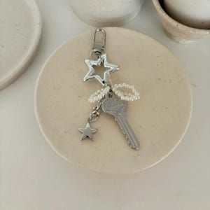 May include: A silver-colored keychain featuring a star and key design. The keychain includes a large star, a small star, and a key charm, all connected with a string of small, white beads. The key has the word "Grace" engraved on it.
