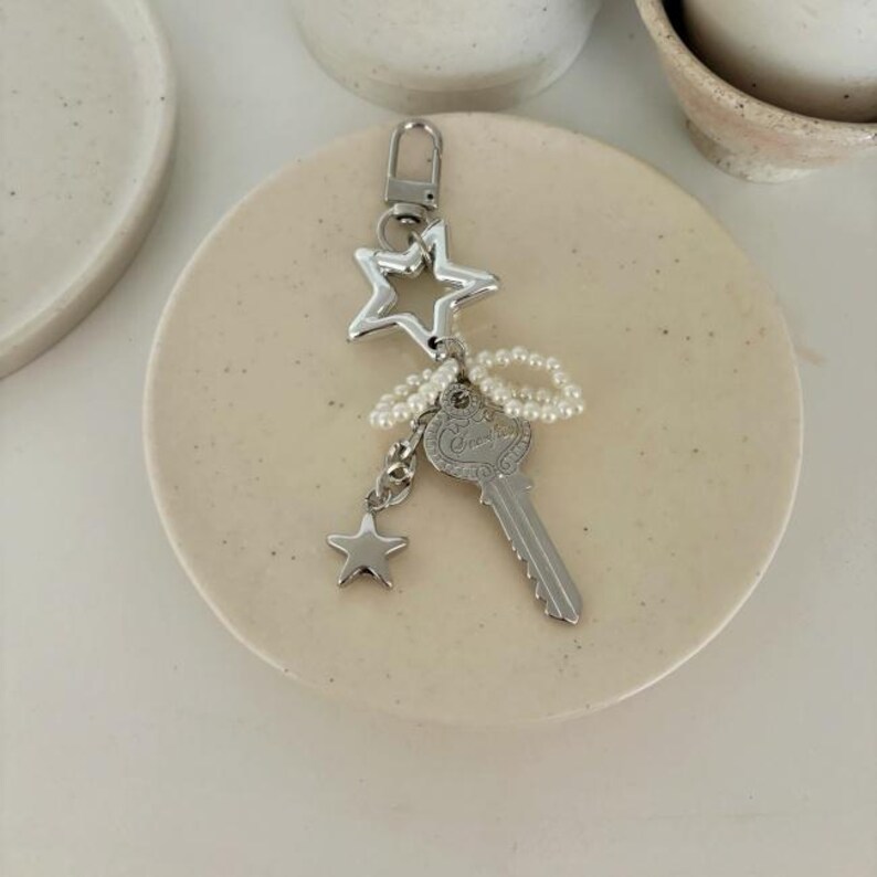 May include: A silver-colored keychain featuring a star and key design. The keychain includes a large star, a small star, and a key charm, all connected with a string of small, white beads. The key has the word "Grace" engraved on it.
