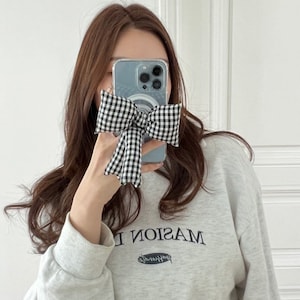 Puede incluir: Una mujer lleva una sudadera gris con la palabra "MASION" impresa en ella. Est&aacute; sosteniendo un lazo a cuadros blanco y negro frente a su cara, cubriendo parcialmente una funda de tel&eacute;fono transparente con un dise&ntilde;o azul y blanco.