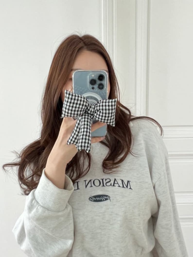 Puede incluir: Una mujer lleva una sudadera gris con la palabra "MASION" impresa en ella. Est&aacute; sosteniendo un lazo a cuadros blanco y negro frente a su cara, cubriendo parcialmente una funda de tel&eacute;fono transparente con un dise&ntilde;o azul y blanco.