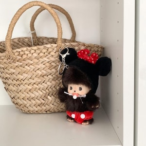 May include: A woven straw tote bag with a pair of handles sits next to a plush toy keychain. The keychain features a small doll wearing a black Mickey Mouse hat with a red bow and red shorts with white polka dots.