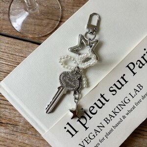 May include: Silver-toned keychain featuring a key, star, and pearl accents. The key has an ornate design, and a small star charm hangs below. The keychain is attached to a clasp and a star-shaped charm. The background includes text that reads "il pleut sur Pari".