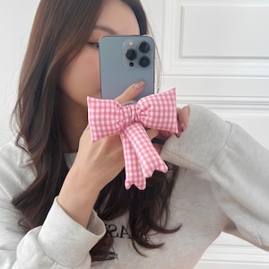 Op de afbeelding: Een roze en witte gingham strik telefoonhouder bevestigd aan een blauwe smartphone. De strik is gemaakt van stof en heeft een geschulpte rand. De telefoon wordt vastgehouden door een persoon die een grijze trui draagt.