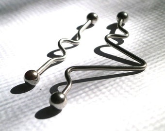 May include: Two silver body jewelry pieces with curved designs. One piece has a wavy design and the other has a more angular design.