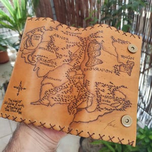 May include: A tan leather wallet featuring a detailed map of Middle Earth, with regions like Gondor and Mordor. The map is etched into the leather, and the wallet has a stitched border and magnetic snap closures. Text on the wallet reads "The West of Middle Earth At The End of The Third Age."