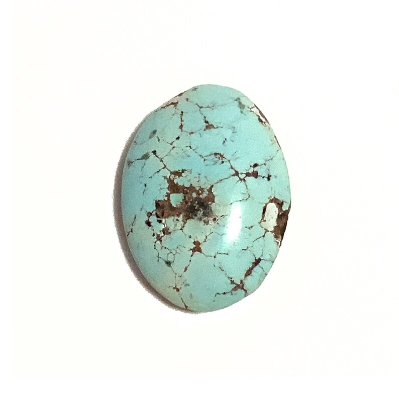 A pale turquoise robins egg blue gemstone with brown matrix markings which is oval in shape. This polished loose turquoise gemstone has a domed rounded top and a flat underside. It is photographed from the top and is on a plain white background.