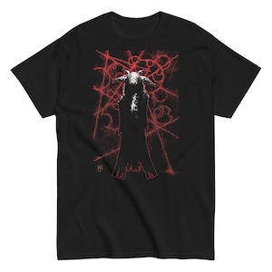 Könnte beinhalten: Schwarzes T-Shirt mit einer rot-weißen Grafik einer gehörnten Figur in einem Umhang, vor einem Hintergrund aus abstrakten roten Linien und Formen. Das Design hat eine abgenutzte, Vintage-Ästhetik.