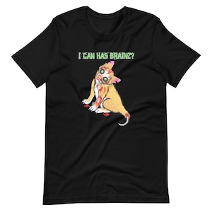 Könnte beinhalten: Schwarzes T-Shirt mit einer Cartoon-Zombie-Katze, die ein Gehirn hält, und dem Text "I CAN HAS BRAINZ?" in Grün.
