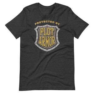 May include: A dark heather grey t-shirt with a graphic of a shield that says "Protected by Plot Armor" in yellow text.
