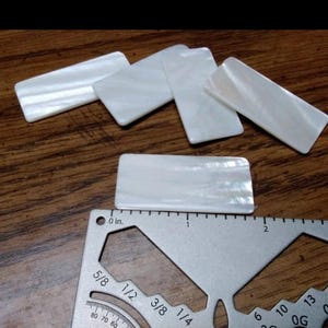 May include: Several rectangular, iridescent white mother-of-pearl pieces are arranged on a wooden surface. A metal ruler is placed near one of the pieces, showing measurements in inches. The mother-of-pearl pieces are approximately 1 inch long.