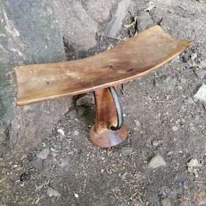 Puede incluir: Un taburete de madera con un asiento curvo y una sola pierna tallada alta. La pierna tiene una correa de cuero negro envuelta alrededor.