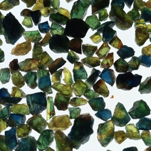 Op de afbeelding: Een verzameling losse groene en blauwe edelstenen. De edelstenen zijn van verschillende vormen en maten en zijn verspreid over een wit oppervlak.