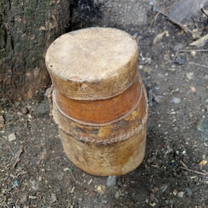 May include: A round, brown leather container with a woven top. The container has a worn, rustic appearance.