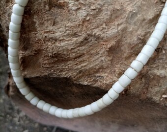 May include: A white beaded necklace made of small, round beads. The necklace is lying on a piece of rough, brown wood.