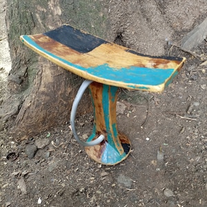 Puede incluir: Un taburete de madera con un asiento curvo y un solo asa. El taburete está pintado en tonos de azul, marrón y negro.