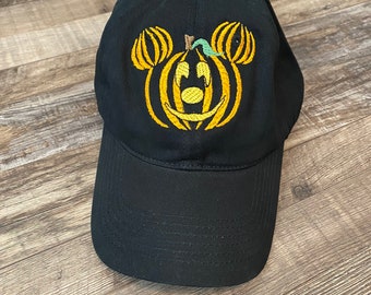 May include: A black baseball cap featuring an embroidered yellow pumpkin design on the front. The pumpkin is shaped like a mouse head with two smaller circular pumpkin ears, a central pumpkin face, and a small green stem at the top center.