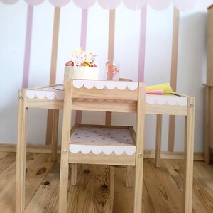 May include: A wooden children's table and chair set with pink and white scalloped trim. The table has a light wood finish and the chair has a pink floral pattern on the seat.