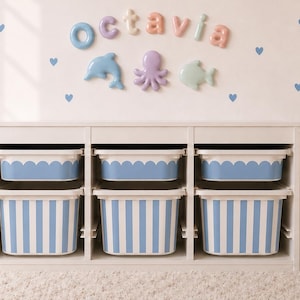 May include: A white storage unit with three sections, each containing two stacked bins. The top bins have scalloped edges, while the bottom bins feature blue and white vertical stripes. The wall displays the name Octavia and sea creature decals.