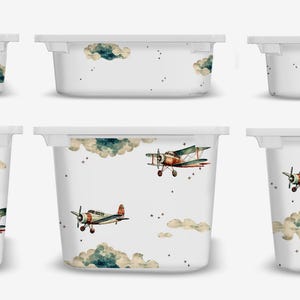 May include: Six white storage bins with lids featuring a vintage airplane and cloud pattern. The airplanes are green, brown, and red with white accents. The clouds are a light blue and gray.