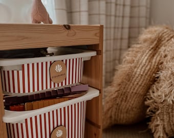 May include: A wooden storage unit with two striped bins, labelled "FIGURINES" and "NURF". A pink felt giraffe figurine sits on top. The bins have red and white vertical stripes. The unit is made of light-coloured wood.