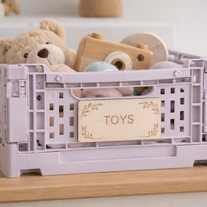 May include: A lilac collapsible storage crate filled with toys. A beige teddy bear, a wooden toy camera, and other toys are visible. A wooden label with the word "TOYS" is attached to the front of the crate.