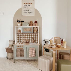 Könnte beinhalten: Ein hellgrünes Spielküchen-Set mit einer braunen und weißen karierten Rückwand, einer braunen Weidenofen-Tür und einem braunen und weißen gestreiften Handtuch, das an der Schranktür hängt. Die Küche ist in einem Spielzimmer mit einem braunen und weißen gestreiften Teppich, einem Holztisch mit einem braunen Schild, auf dem "Getränke" steht, und zwei Plüschtieren, einem braunen Bären und einem schwarz-weißen Panda, eingerichtet.
