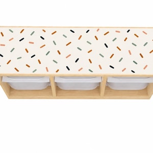 Puede incluir: Una unidad de almacenamiento rectangular de madera con tres compartimentos abiertos, cada uno con un contenedor de plástico blanco. La superficie superior tiene un acabado en color blanco roto y presenta un patrón repetitivo de pequeñas rayas dispersas en terracota, verde salvia y azul marino oscuro.