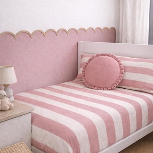 Può includere: Una camera da letto con un letto bianco, con un copripiumino e un cuscino a righe rosa e bianche. Un cuscino rotondo rosa con un bordo a balze è sopra. La parete è dipinta di rosa con un bordo in legno smerlato.
