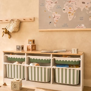 May include: A white wooden storage unit with three sections, each containing two stacked plastic bins. The top bins have a scalloped green edge, while the bottom bins feature vertical green and white stripes. Wooden blocks with numbers and animal illustrations are placed on top.
