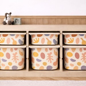 Könnte beinhalten: Ein helles Holzregal mit sechs Fächern, in denen jeweils eine gemusterte Box steht. Die Boxen haben einen cremefarbenen Hintergrund mit bunten abstrakten Formen in Orange, Gelb, Rosa und Lila. Ein Stofftier und kleine Dekorationsartikel stehen obenauf.