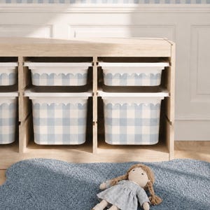 May include: A wooden storage unit with six cubbies, each holding a white bin with a scalloped edge and a blue gingham pattern. A blue rug and a doll in a blue dress are in front of the unit. A blue and white scalloped border is on the wall.