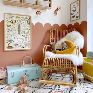 Op de afbeelding: Een lichtblauwe metalen opbergkist met een schommelstoel op de achtergrond. De schommelstoel is gemaakt van natuurlijke wicker en heeft een witte nep bontdeken en een bruin en geel kussen met een berenkop erop. De muur achter de stoel is terracotta geverfd met een gegolfde rand.