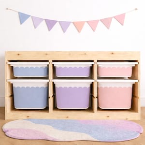 May include: A wooden storage unit with six pastel-colored bins. The bins are blue, purple, and pink with white scalloped edges. A matching rug and a pastel banner are in the foreground and background, respectively. This storage unit is ideal for a child's room.