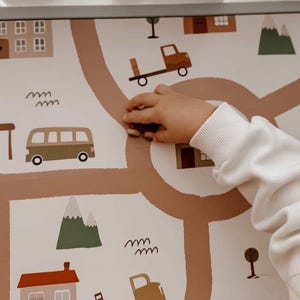 May include: A children's play mat featuring a road map design with illustrations of houses, mountains, trees, and vehicles in shades of brown, green, and beige. A hand is interacting with the mat, possibly placing a toy truck on the road.