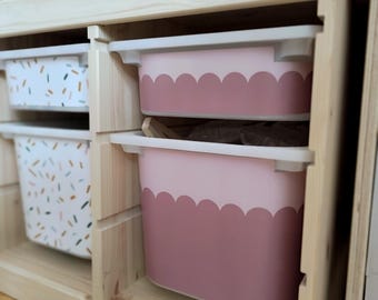 May include: A wooden storage unit with two pink storage bins with scalloped edges. The bins are in a light wood cabinet with a white top.