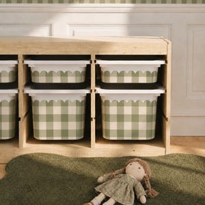 May include: Wooden storage unit with six compartments, each holding a white and green gingham patterned bin. A green rug and a doll with a floral dress are in front of the unit. The wall has a scalloped border.