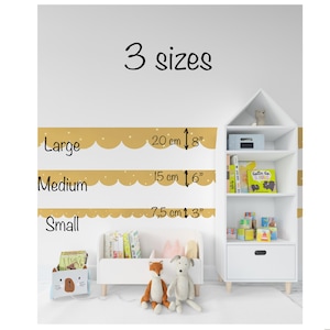 Puede incluir: Una pared blanca con bordes dorados festoneados que indican tres tamaños: Grande (20 cm), Mediano (15 cm) y Pequeño (7,5 cm). El texto "3 tamaños" está en la parte superior. Una estantería blanca y un pequeño banco blanco con juguetes y peluches están en primer plano.