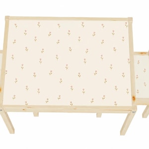 Puede incluir: Un juego de mesa y dos sillas para niños. La mesa y las sillas son de madera clara. Los asientos de la mesa y las sillas tienen una superficie color crema con un patrón repetido de pequeñas flores rosas. El conjunto está diseñado para niños.