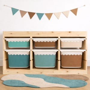 May include: A wooden storage unit with six rectangular bins in shades of teal, brown, and beige, each with a scalloped white trim. A matching rug and a banner of triangular flags in similar colors are in the foreground and background.