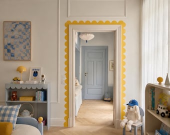 Op de afbeelding: Een kamerinterieur met een deuropening omlijst door een gele geschulpte rand. De ruimte bevat een lichtblauwe boekenplank met een geschulpte bovenkant, een bed met een blauw frame, een wit kleed met blauwe streepjes en een lichtblauwe deur.