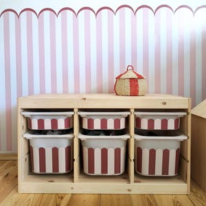 May include: A wooden storage unit with six compartments, each holding a white and red striped bin. A woven basket with a red and white striped lid sits on top. The background features a pink and white striped wall.