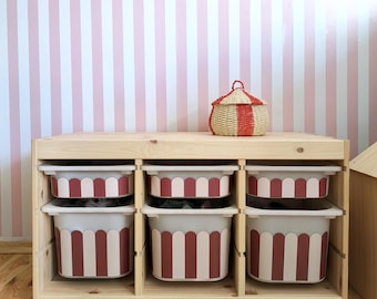 May include: A wooden storage unit with six compartments, each holding a white and red striped bin. A woven basket with a red and white striped lid sits on top. The background features a pink and white striped wall.