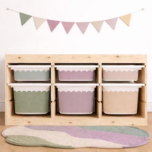 May include: A wooden storage unit with six pastel-colored bins. The bins are in shades of green, purple, and tan, each with a white scalloped edge. A colorful banner hangs above the unit, and a patterned rug is in front.