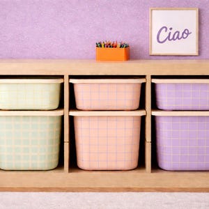 May include: A wooden storage unit with six pastel-colored bins, each with a grid pattern. An orange container holds colored pencils, and a framed sign reads "Ciao". The bins are in shades of green, peach, and purple.