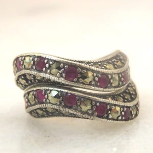 以下が含まれることがあります： 渦巻き模様のシルバーリング。リングには、小さな丸い赤い宝石と、より小さな丸いシルバー色の石が交互に並んでいます。リングは白い背景に配置されています。