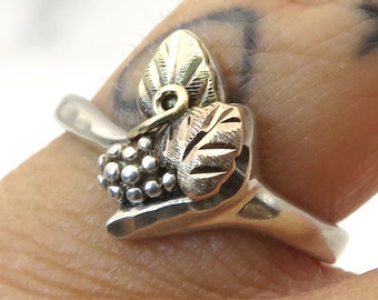 Puede incluir: Un anillo de plata con un diseño de hoja de oro y un racimo de pequeñas cuentas de plata. El anillo se lleva en un dedo.