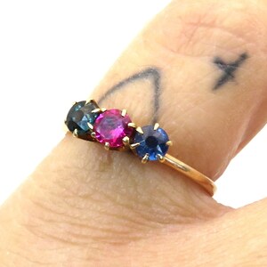 May include: A gold ring with three gemstones: a dark blue, a pink, and a light blue. The ring is worn on a finger with a tattoo.
