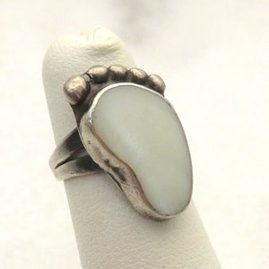 May include: A silver ring with a white mother of pearl stone in the shape of a foot. The ring is set with small silver beads around the stone.