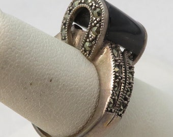 May include: A silver ring with a black onyx stone and marcasite accents. The ring is on a white ring display stand.