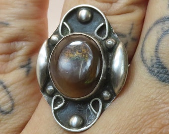 Op de afbeelding: Een zilveren ring met een ovale, iriserende edelsteen gezet in een gedetailleerd ontwerp. De ring zit aan een vinger met tatoeages.